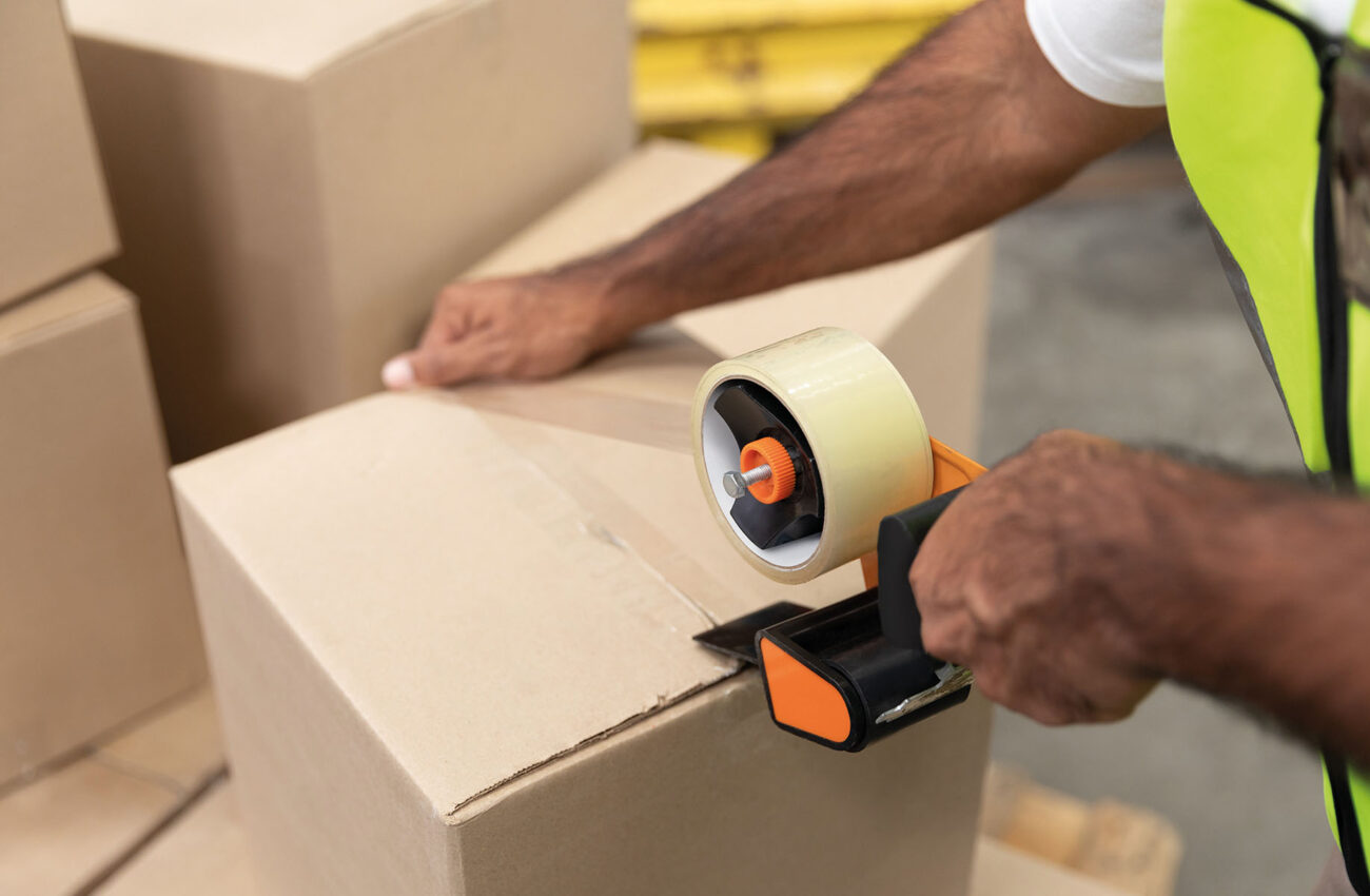 Male worker packing cardboard box with tape gun dispenser in war